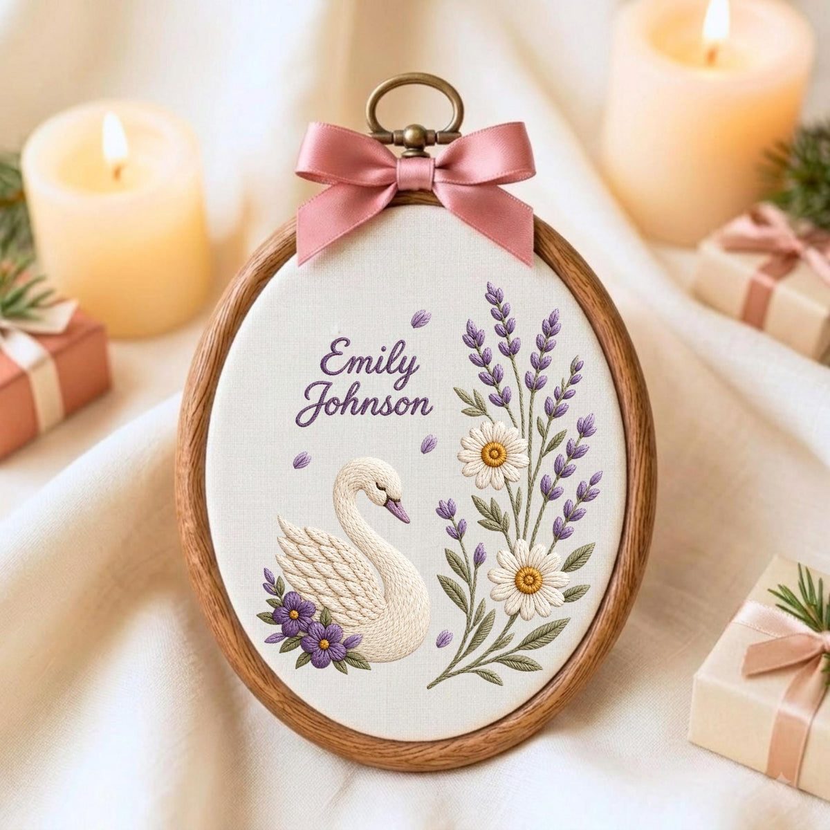 May include: An oval wooden embroidery hoop containing white fabric with a hand-stitched design of a white swan, purple flowers, lavender sprigs, and white daisies. The text Emily Johnson is embroidered in purple. A pink satin bow sits at the top.