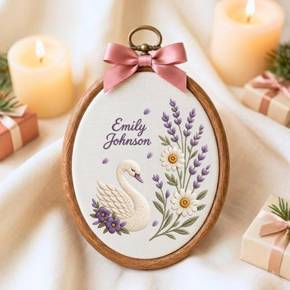 May include: An oval wooden embroidery hoop containing white fabric with a hand-stitched design of a white swan, purple flowers, lavender sprigs, and white daisies. The text Emily Johnson is embroidered in purple. A pink satin bow sits at the top.