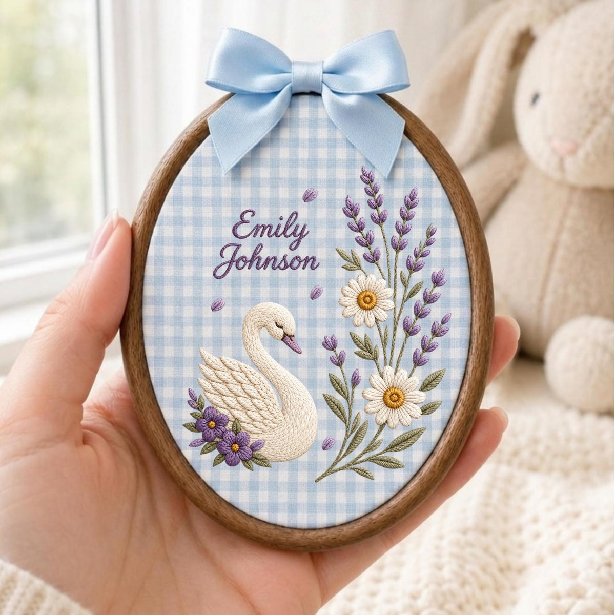 May include: An oval wooden embroidery hoop featuring a light blue and white gingham fabric background. The embroidery depicts a white swan, purple flowers, lavender sprigs, and two daisies. The text Emily Johnson is stitched in purple script.