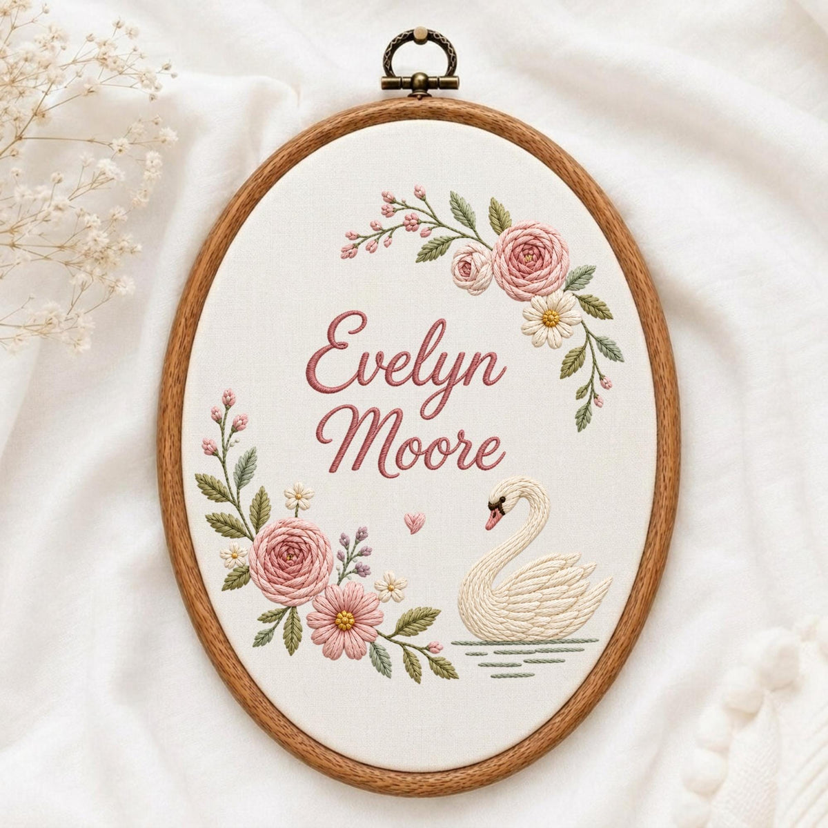 May include: An oval wooden embroidery hoop containing white fabric with the embroidered name Evelyn Moore in pink script. The design features pink roses, green leaves, small white flowers, a small pink heart, and a white swan on water.