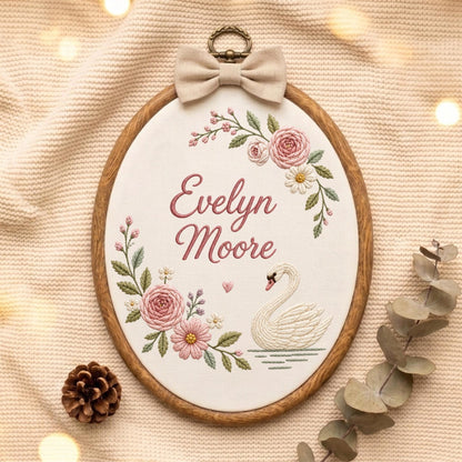 May include: An oval wooden embroidery hoop containing white fabric with the embroidered name Evelyn Moore in pink script. The design features a white swan on water, pink roses, small white flowers, green leaves, and a beige fabric bow at the top.