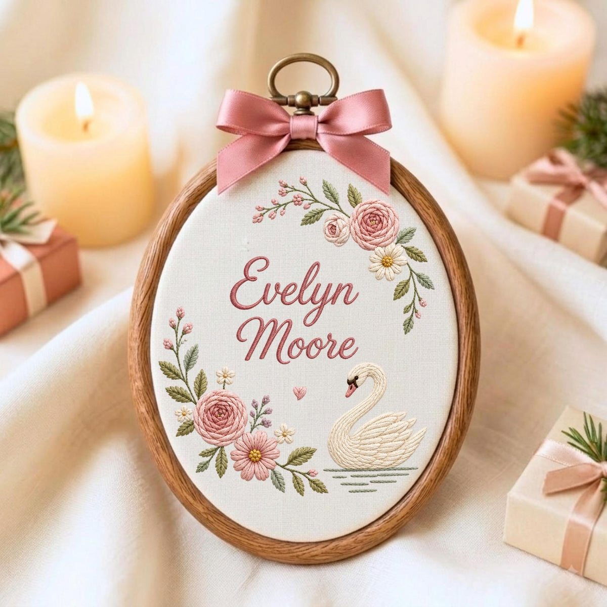 May include: An oval wooden embroidery hoop containing white fabric with the embroidered name Evelyn Moore in pink script. The design features a white swan, pink and white flowers, and green leaves. A pink satin ribbon bow is attached at the top.