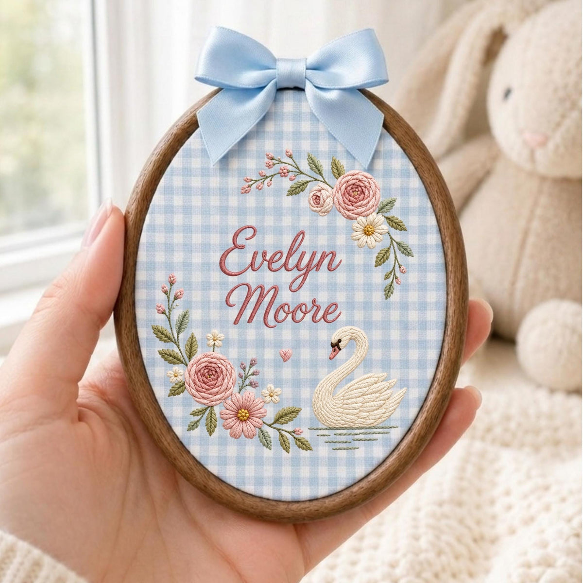 May include: An oval wooden embroidery hoop containing fabric with a light blue and white gingham pattern. The fabric features embroidered pink roses, green leaves, a white swan, and the text Evelyn Moore in pink, topped with a light blue satin bow.