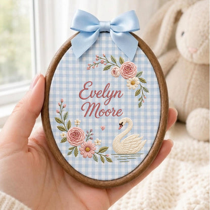 May include: An oval wooden embroidery hoop containing fabric with a light blue and white gingham pattern. The fabric features embroidered pink roses, green leaves, a white swan, and the text Evelyn Moore in pink, topped with a light blue satin bow.