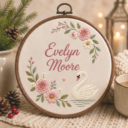 May include: A circular wooden embroidery hoop containing cream-colored fabric with the embroidered name Evelyn Moore in pink script. The design features a white swan swimming on water, surrounded by pink roses, small white flowers, and green leaves.