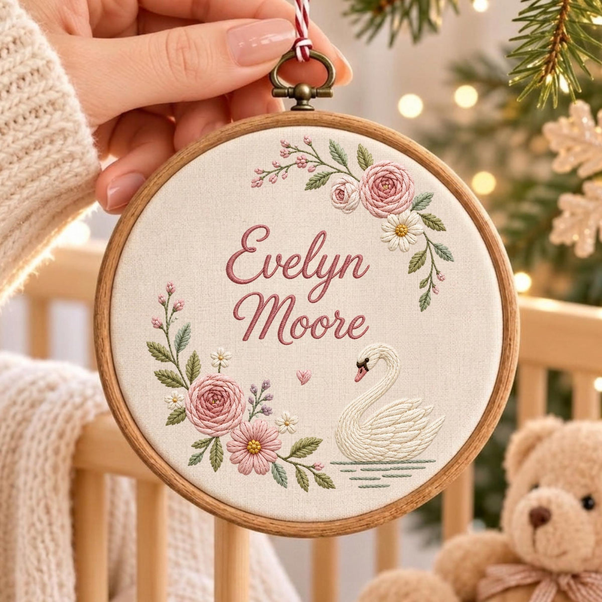 May include: A circular wooden embroidery hoop containing off-white fabric with the embroidered name Evelyn Moore in pink script. The design features a white swan, pink roses, and green leaves arranged in a floral wreath around the text.