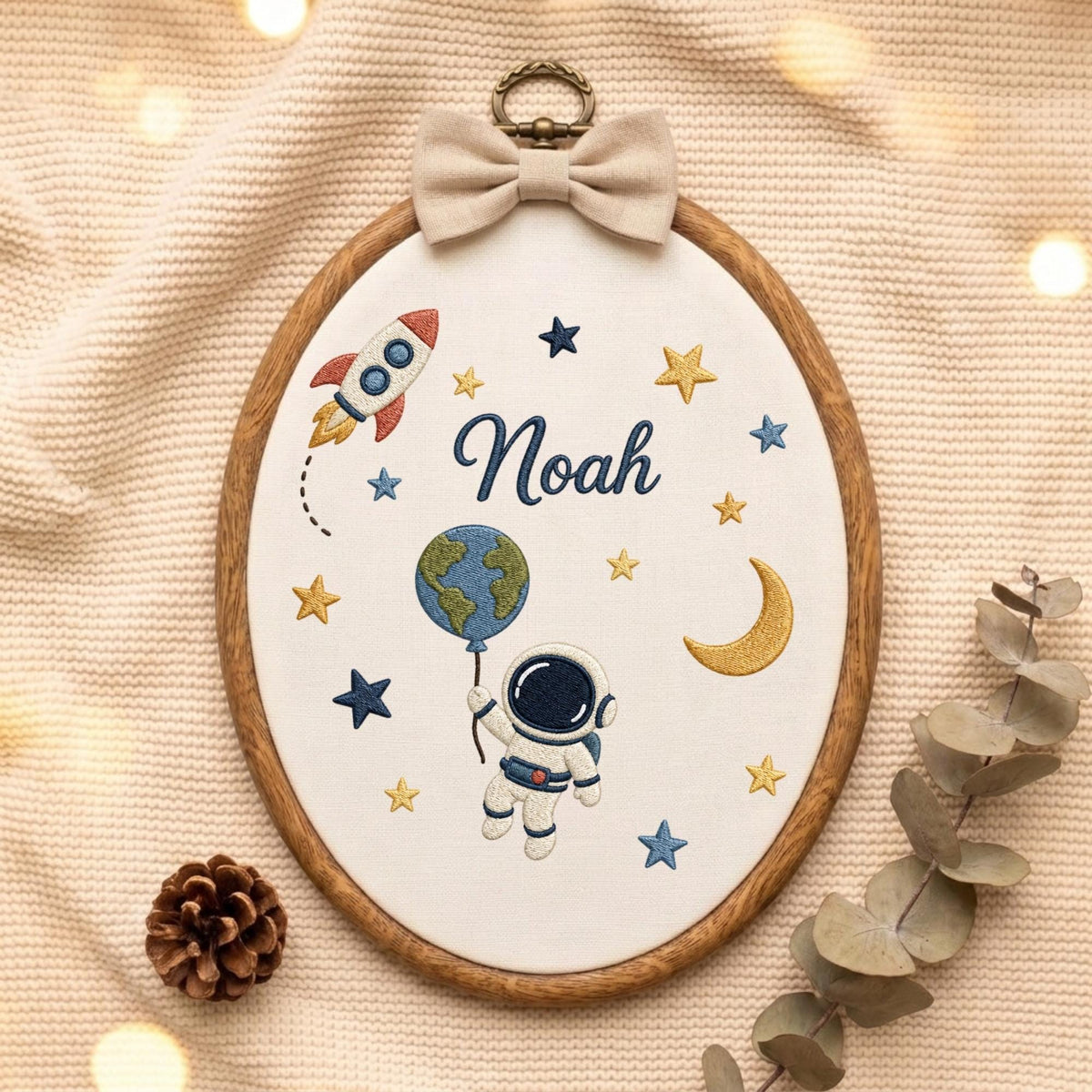 May include: An oval embroidery hoop featuring a white fabric background with a space-themed design. The embroidery includes a rocket, a crescent moon, stars, an astronaut holding a globe balloon, and the name Noah in dark blue cursive script.