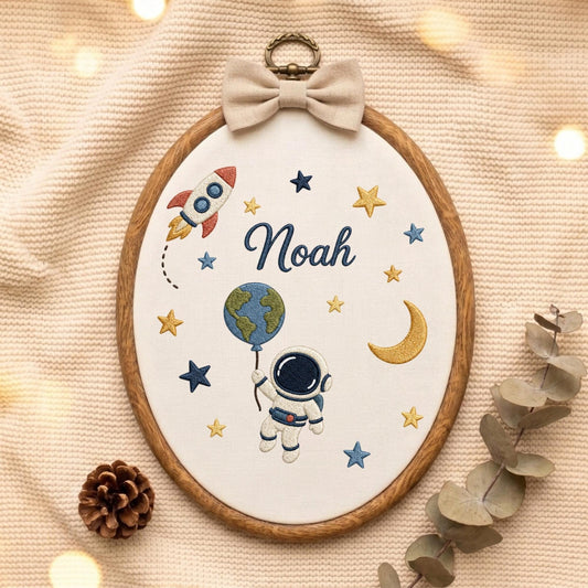 May include: An oval embroidery hoop featuring a white fabric background with a space-themed design. The embroidery includes a rocket, a crescent moon, stars, an astronaut holding a globe balloon, and the name Noah in dark blue cursive script.