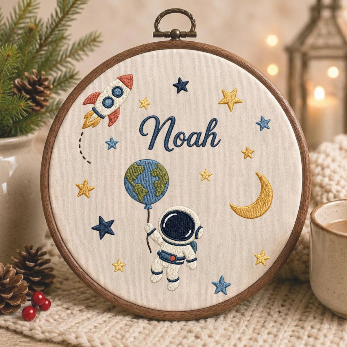 May include: A circular wooden embroidery hoop containing off-white fabric with a space-themed design. The embroidery features a rocket, a crescent moon, stars, a planet Earth balloon, and an astronaut. The name Noah is stitched in blue.