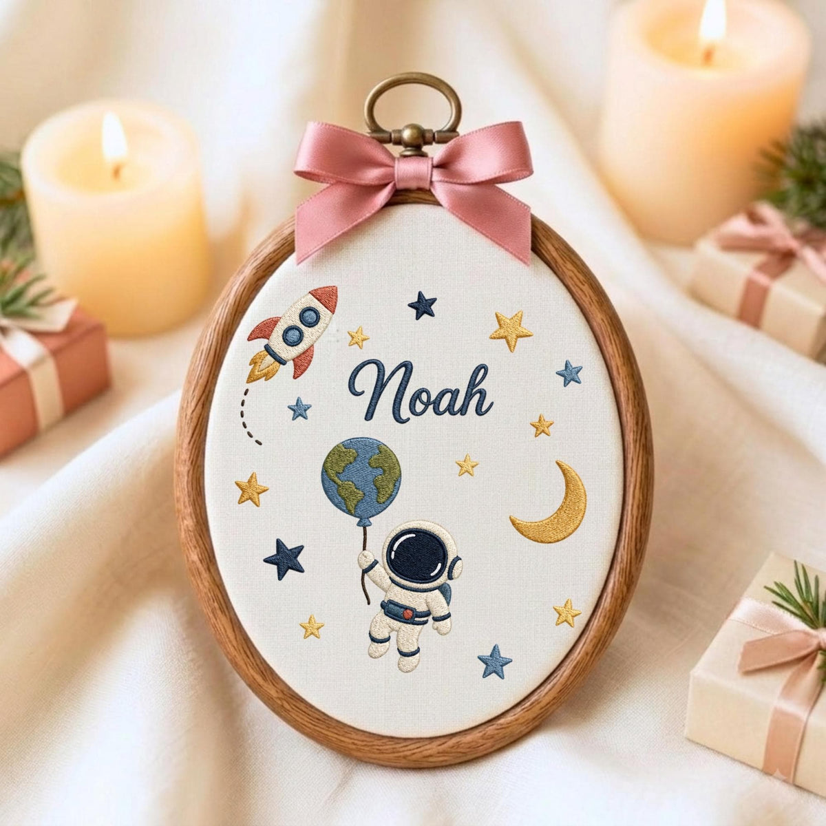 May include: An oval embroidery hoop featuring a white fabric background with the name Noah stitched in dark blue. The design includes a colorful rocket, a crescent moon, yellow and blue stars, and an astronaut holding a globe balloon.