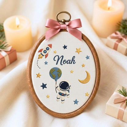 May include: An oval embroidery hoop featuring a white fabric background with the name Noah stitched in dark blue. The design includes a colorful rocket, a crescent moon, yellow and blue stars, and an astronaut holding a globe balloon.