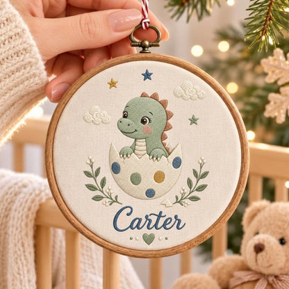 May include: A circular wooden embroidery hoop containing a cream-colored fabric panel. The fabric features an embroidered green dinosaur hatching from a spotted egg, surrounded by small stars, clouds, and leaves. The name Carter is stitched in blue.