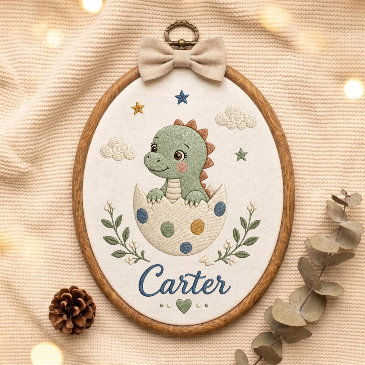 May include: An oval embroidery hoop featuring a green dinosaur hatching from a white egg with blue and yellow polka dots. The design includes embroidered clouds, stars, green leafy branches, and the name Carter in blue script with a small green heart.