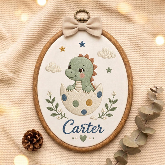 May include: An oval embroidery hoop featuring a green dinosaur hatching from a white egg with blue and yellow polka dots. The design includes embroidered clouds, stars, green leafy branches, and the name Carter in blue script with a small green heart.