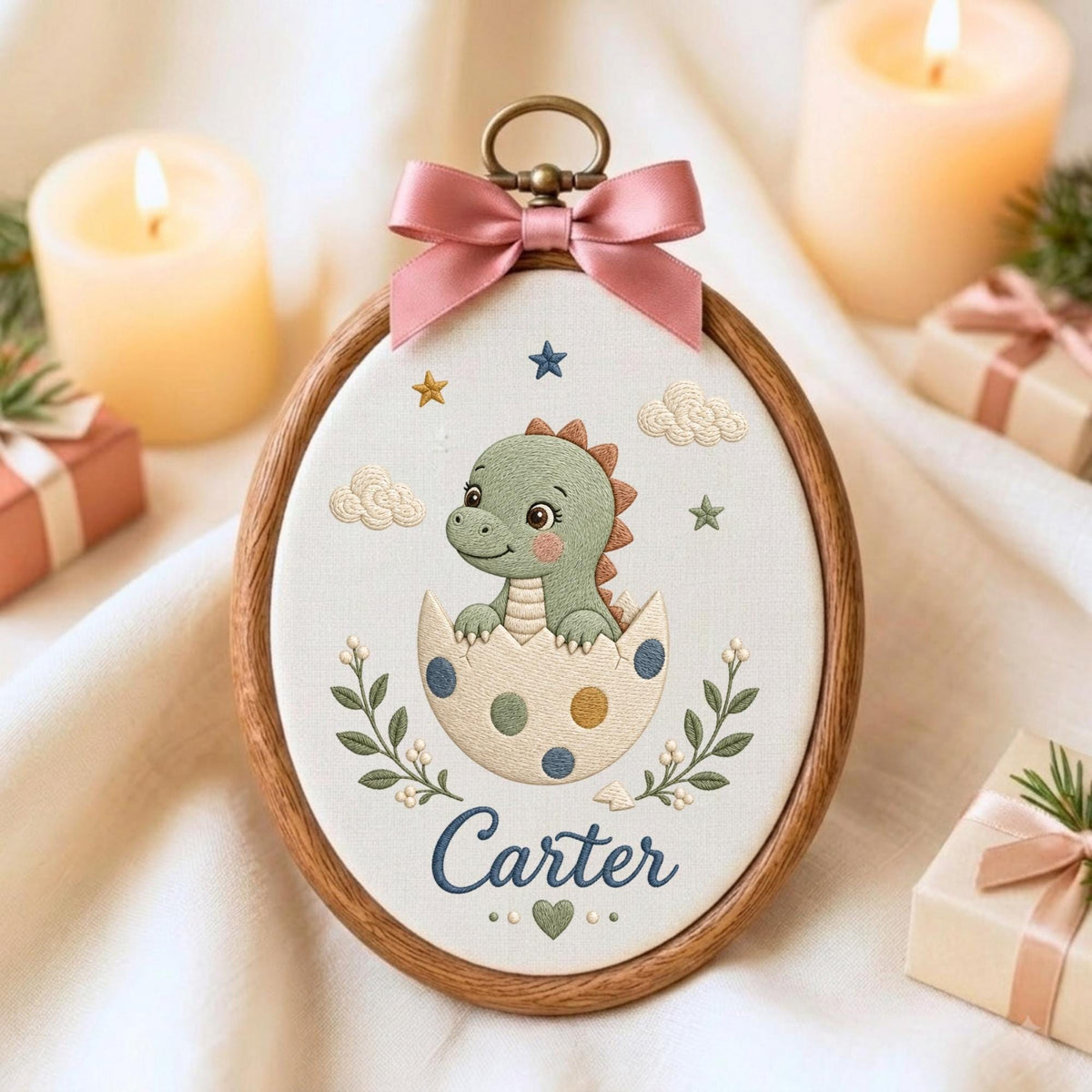 May include: An oval wooden embroidery hoop containing white fabric with a stitched green dinosaur hatching from a polka-dot egg. The design includes small stars, clouds, green leafy branches, a heart, and the name Carter in blue script text.