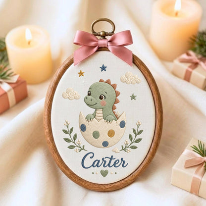 May include: An oval wooden embroidery hoop containing white fabric with a stitched green dinosaur hatching from a polka-dot egg. The design includes small stars, clouds, green leafy branches, a heart, and the name Carter in blue script text.