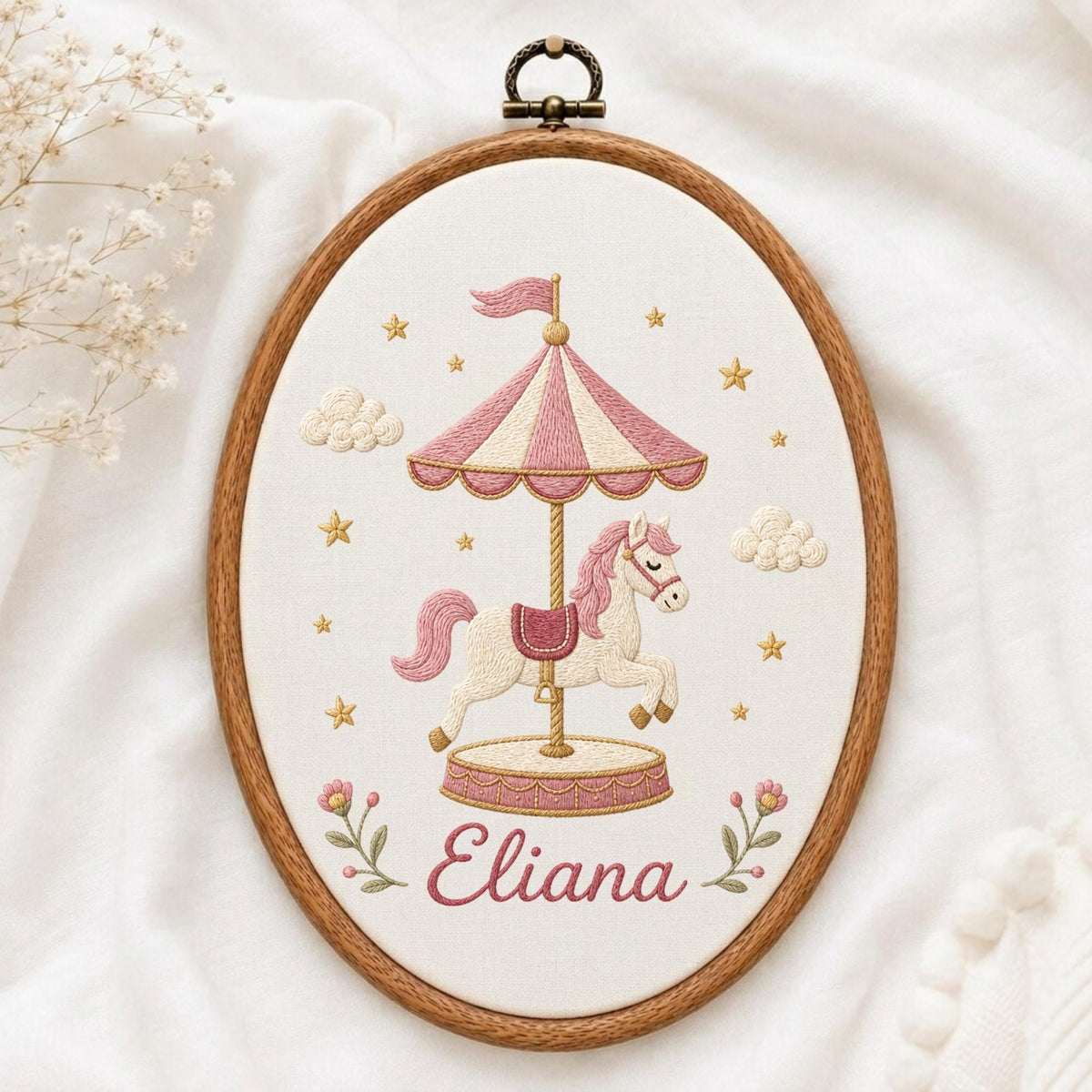 May include: An oval embroidery hoop containing a white fabric panel with a stitched carousel horse design. The carousel features a pink and white striped canopy, a cream-colored horse, gold stars, clouds, and the name Eliana in pink script.