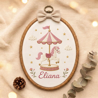 May include: An oval embroidery hoop featuring a carousel horse design stitched on white fabric. The carousel has pink and white stripes, and the horse is white with a pink mane. The name Eliana is embroidered in pink script below the carousel.