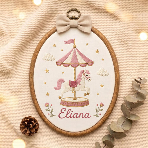 May include: An oval embroidery hoop featuring a carousel horse design stitched on white fabric. The carousel has pink and white stripes, and the horse is white with a pink mane. The name Eliana is embroidered in pink script below the carousel.
