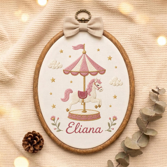 May include: An oval embroidery hoop featuring a carousel horse design stitched on white fabric. The carousel has pink and white stripes, and the horse is white with a pink mane. The name Eliana is embroidered in pink script below the carousel.