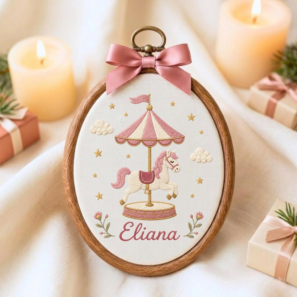 May include: An oval wooden embroidery hoop containing white fabric with a stitched carousel horse design in pink and cream. The carousel features a striped canopy, a pink saddle, and the name Eliana in cursive script. A pink satin bow sits at the top.