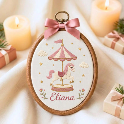 May include: An oval wooden embroidery hoop containing white fabric with a stitched carousel horse design in pink and cream. The carousel features a striped canopy, a pink saddle, and the name Eliana in cursive script. A pink satin bow sits at the top.
