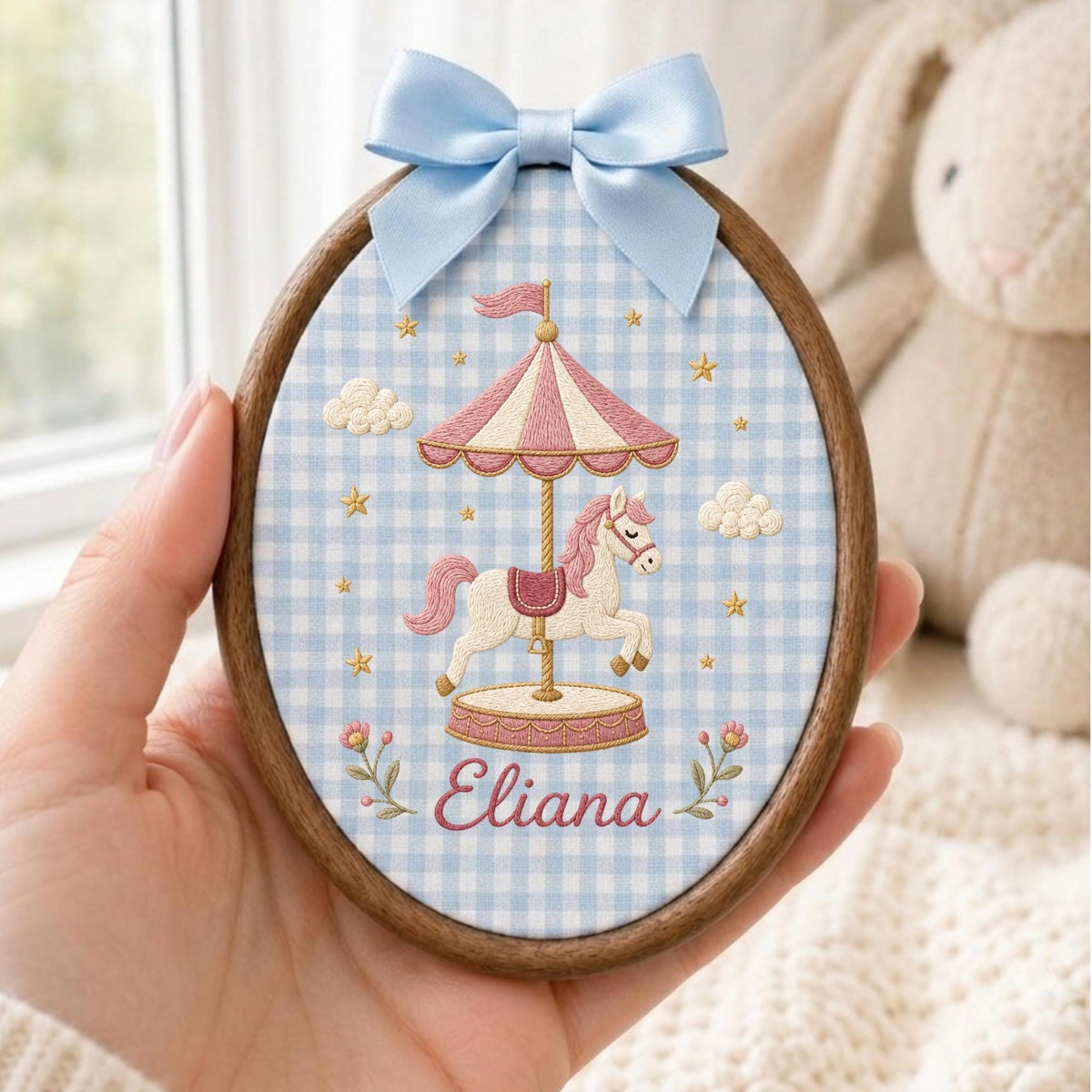 May include: An oval wooden embroidery hoop featuring a light blue and white gingham fabric background. The embroidery depicts a carousel horse under a pink and white striped canopy, surrounded by clouds, stars, and the name Eliana in pink script.