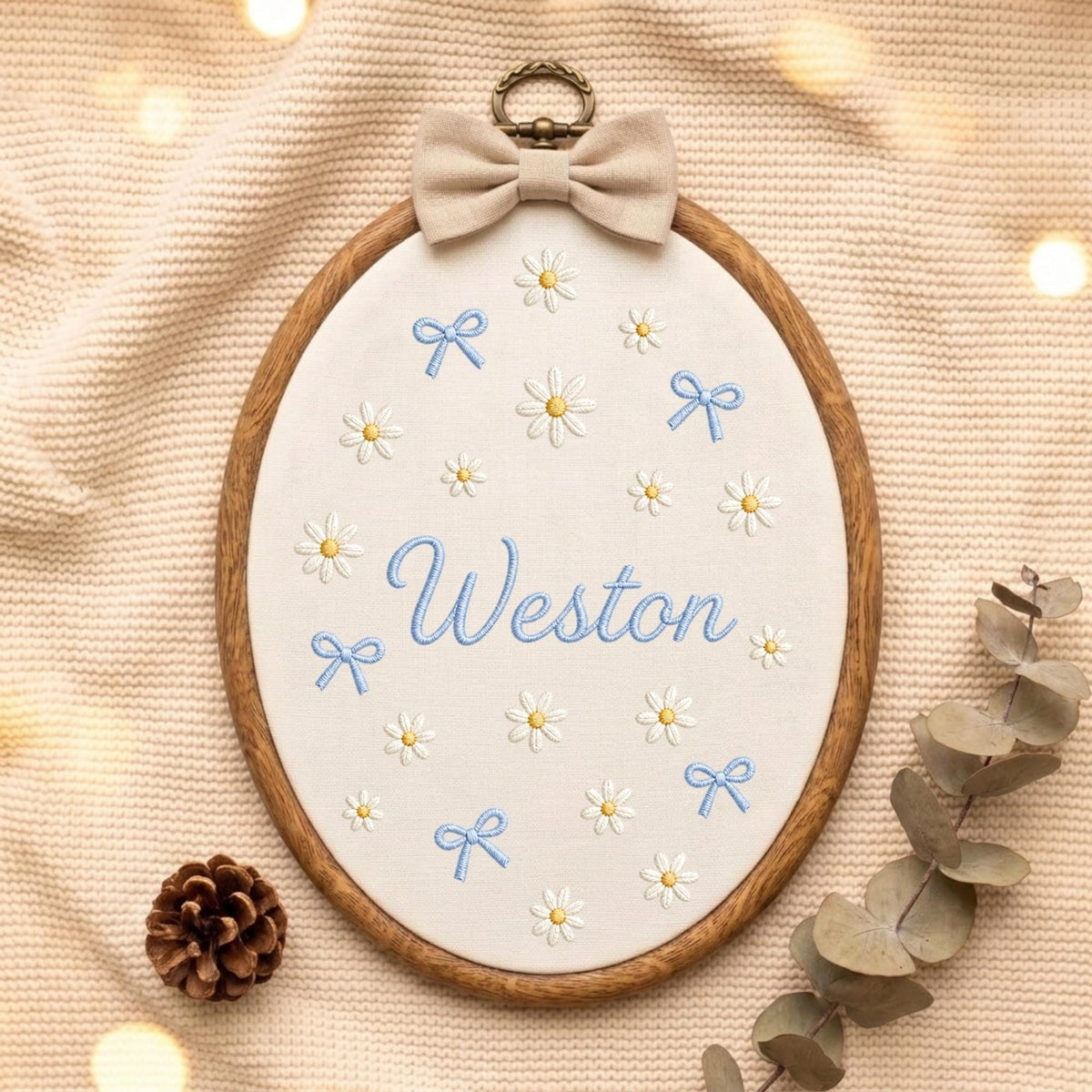 May include: An oval wooden embroidery hoop containing white fabric with the embroidered name Weston in blue script, surrounded by small white daisy flowers with yellow centers and light blue bows, topped with a beige fabric bow.