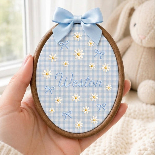 May include: An oval wooden embroidery hoop containing light blue and white gingham fabric. The fabric features embroidered white daisies, small blue bows, and the name Weston in blue script. A light blue satin ribbon bow is attached at the top.