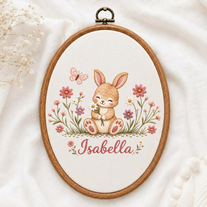 May include: An oval wooden embroidery hoop containing a fabric panel with a stitched illustration of a light brown bunny holding a white daisy. The design includes pink flowers, green stems, a small pink butterfly, and the name Isabella in pink text.