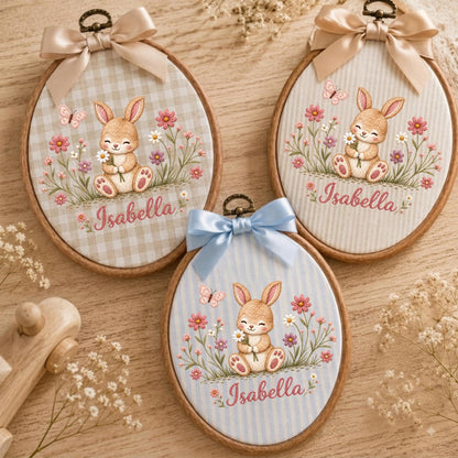May include: Three oval wooden embroidery hoops featuring fabric backgrounds with a bunny, flowers, and a butterfly. Two hoops have beige ribbons and one has a blue ribbon. Each hoop displays the name Isabella in cursive script text.