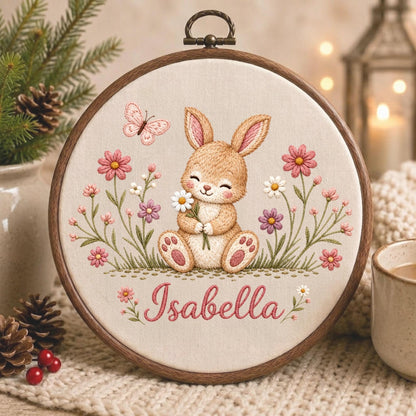 May include: A circular wooden embroidery hoop containing a fabric panel with a stitched design of a light brown bunny holding a white daisy. The scene includes pink and purple flowers, green stems, and a pink butterfly. The name Isabella is embroidered in pink script below the bunny.