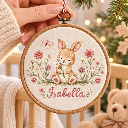 May include: A circular wooden embroidery hoop containing a cream-colored fabric panel. The fabric features an embroidered design of a brown bunny holding a white daisy, surrounded by pink and purple flowers, a butterfly, and the name Isabella.