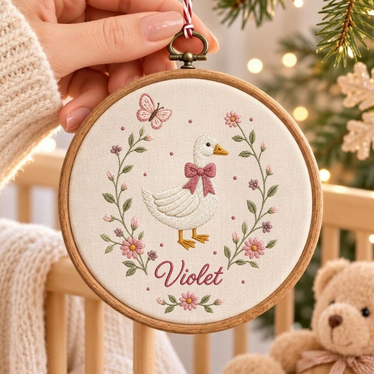 May include: A circular wooden embroidery hoop containing a white fabric panel with a hand-stitched design of a white goose wearing a pink bow. The goose is surrounded by a floral wreath with pink flowers, green leaves, and a butterfly. The name Violet is stitched in pink script below the goose.