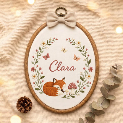 May include: An oval wooden embroidery hoop containing white fabric with a hand-stitched design. The embroidery features a sleeping orange fox, a mushroom, butterflies, and floral vines surrounding the name Clara in pink script, topped with a beige bow.