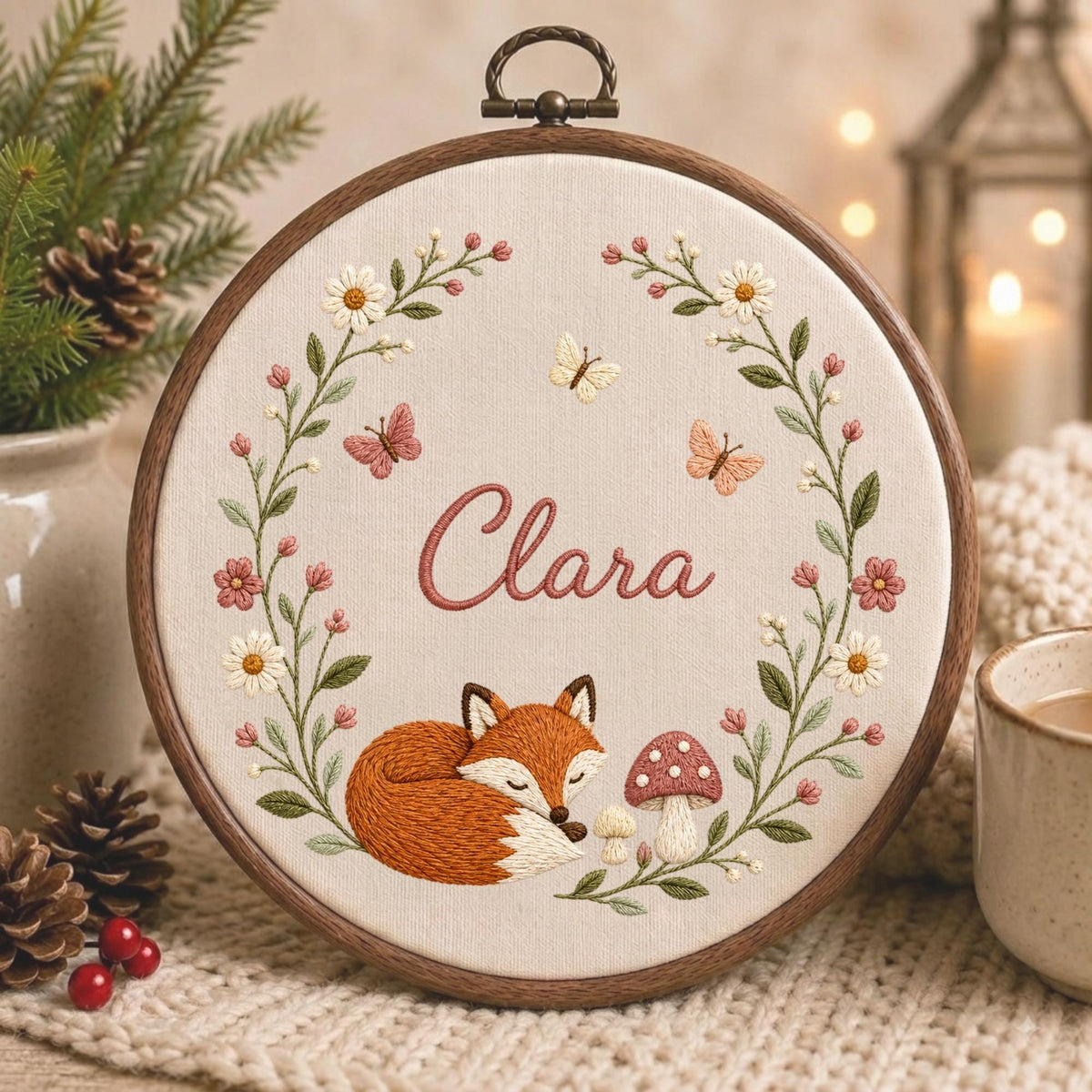 May include: A circular wooden embroidery hoop containing a cream-colored fabric with a hand-stitched design. The embroidery features a sleeping orange fox, a mushroom, butterflies, and a floral wreath surrounding the name Clara in pink script.