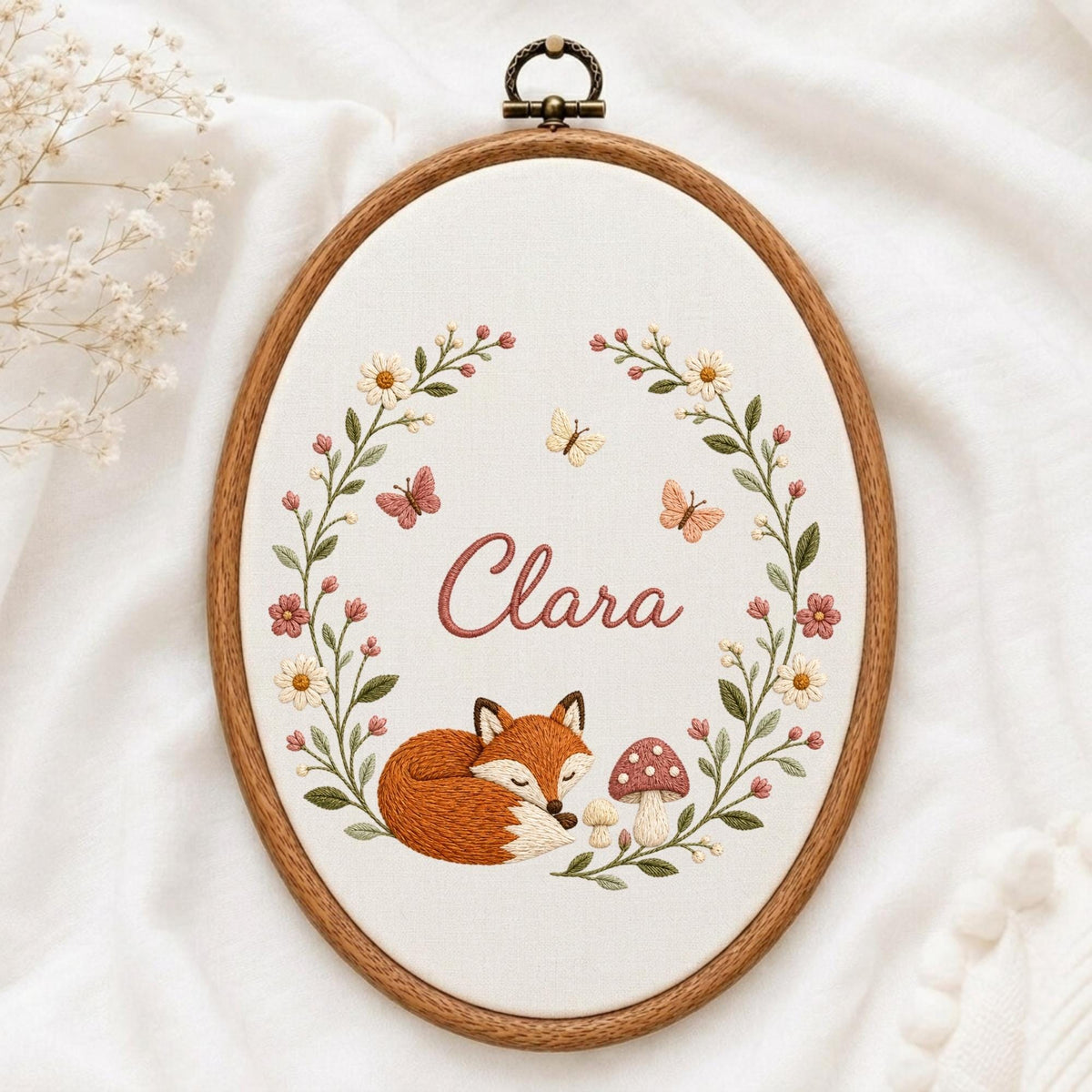 May include: An oval wooden embroidery hoop containing white fabric with a hand-stitched design. The embroidery features a sleeping orange fox, a mushroom, and a floral wreath with butterflies. The name Clara is stitched in pink cursive text.