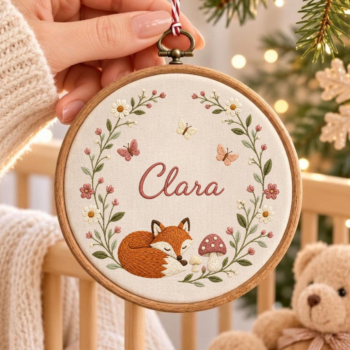 May include: A circular wooden embroidery hoop containing a cream-colored fabric panel. The fabric features a hand-stitched design of a sleeping orange fox, a mushroom, and a floral wreath with butterflies. The name Clara is embroidered in pink.
