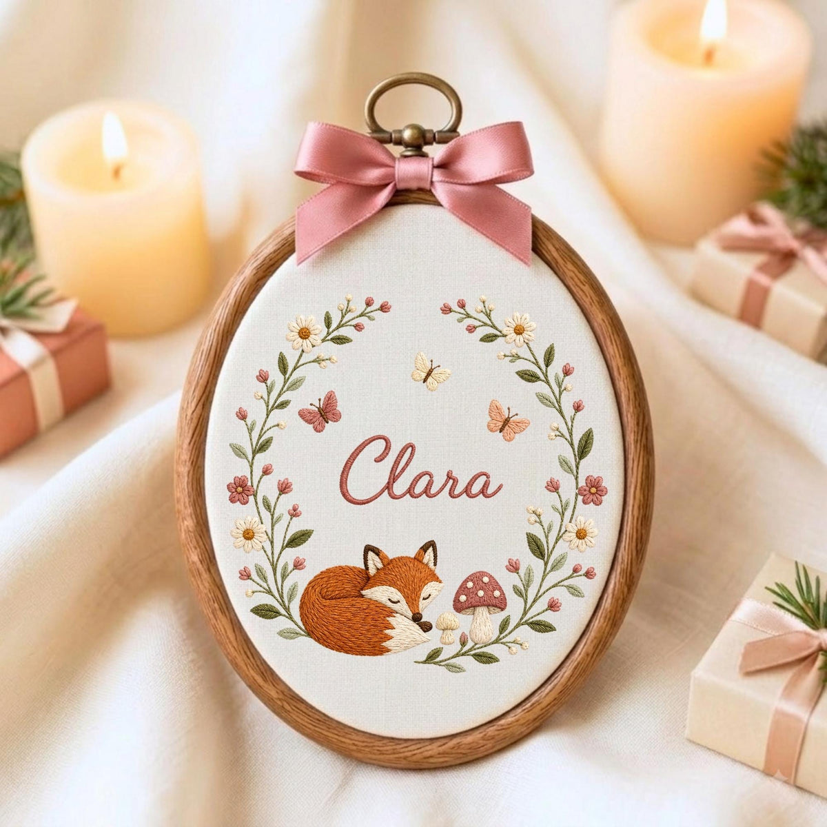May include: An oval wooden embroidery hoop containing white fabric with a hand-stitched design of a sleeping fox, a mushroom, flowers, and butterflies. The name Clara is embroidered in the center in pink script, topped with a pink satin bow.