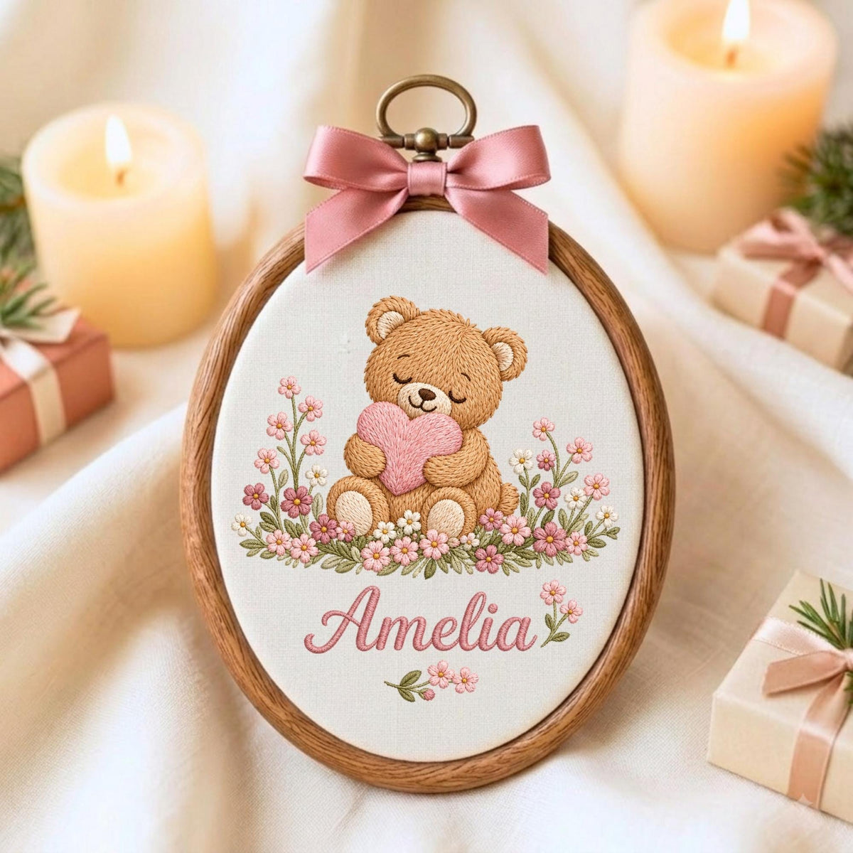 May include: An oval wooden embroidery hoop containing a fabric panel with a stitched illustration of a brown teddy bear holding a pink heart, surrounded by small pink and white flowers. The name Amelia is embroidered below in pink script.