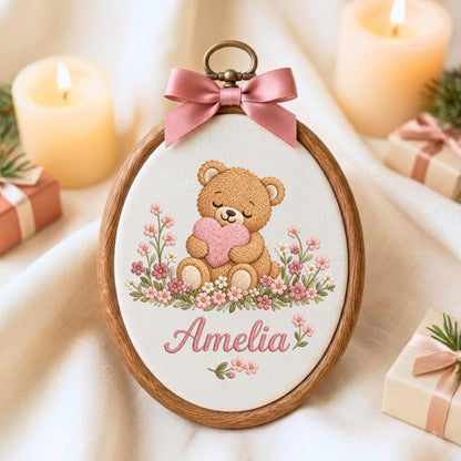 May include: An oval wooden embroidery hoop containing a fabric panel with a stitched illustration of a brown teddy bear holding a pink heart, surrounded by small pink and white flowers. The name Amelia is embroidered below in pink script.