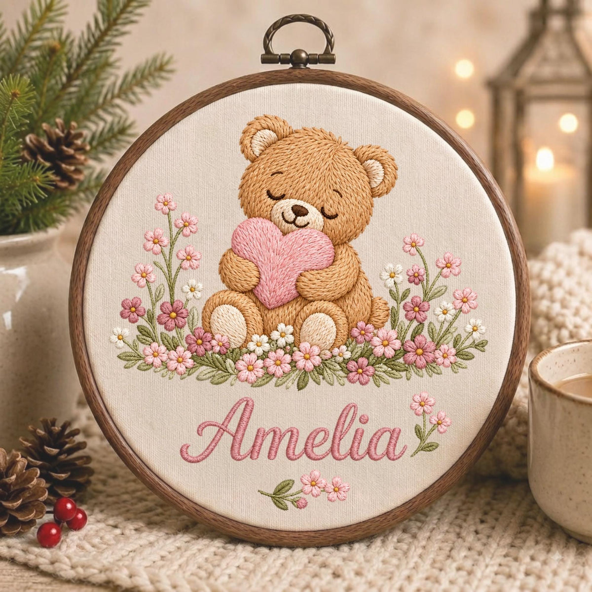 May include: A circular wooden embroidery hoop containing a fabric panel with a stitched design of a brown teddy bear holding a pink heart. The bear is surrounded by pink and white flowers, with the name Amelia embroidered in pink script below.
