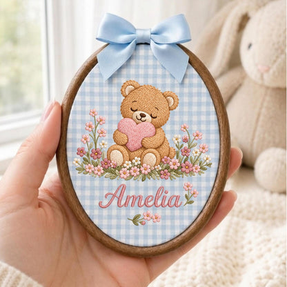 May include: An oval wooden embroidery hoop frame containing fabric with a light blue gingham pattern. The fabric features an embroidered brown teddy bear holding a pink heart, surrounded by small pink and white flowers, with the name Amelia in pink.