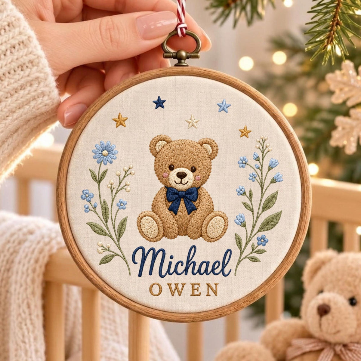 May include: A circular wooden embroidery hoop containing a cream-colored fabric with a stitched design of a brown teddy bear wearing a dark blue bow tie. The bear is flanked by blue floral sprigs and small stars, with the name Michael Owen embroidered below.