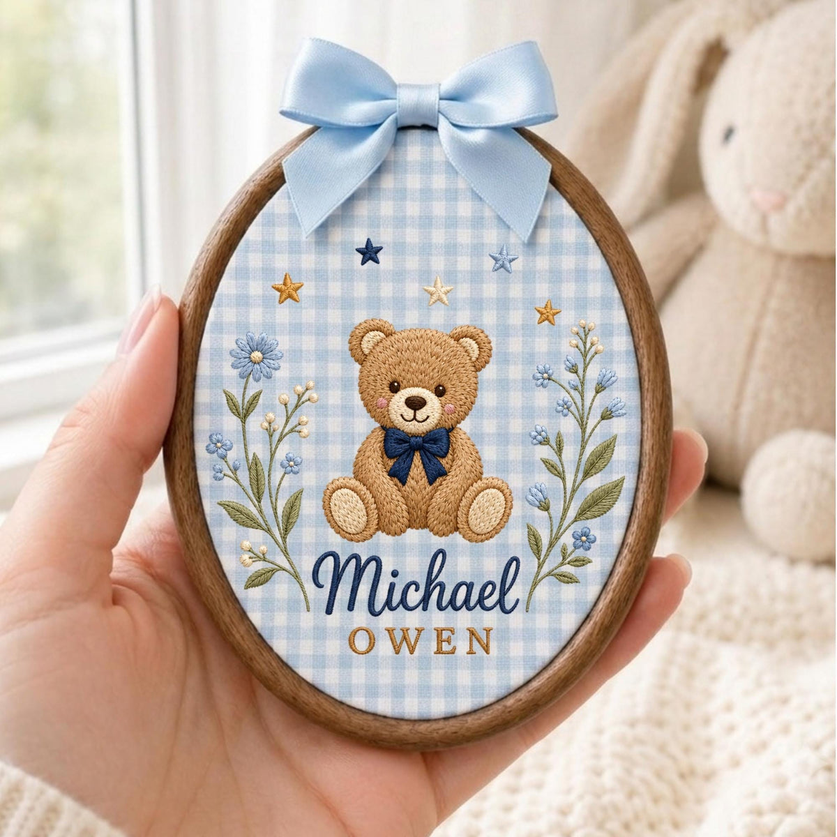 May include: An oval wooden embroidery hoop containing light blue and white gingham fabric. A brown teddy bear with a dark blue bow tie is centered, flanked by blue floral stems and small stars. The text Michael OWEN is embroidered below.