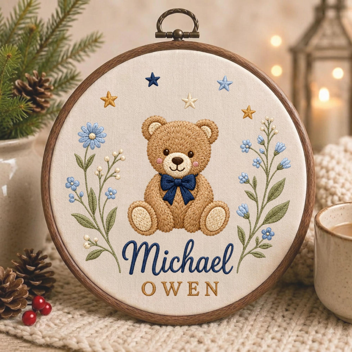 May include: A circular wooden embroidery hoop containing a cream-colored fabric panel. The embroidery features a central brown teddy bear with a dark blue bow tie, flanked by blue floral sprigs and stars. The text reads Michael in blue and OWEN in gold.