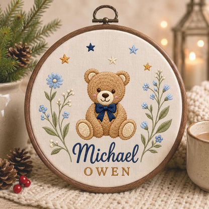 May include: A circular wooden embroidery hoop containing a cream-colored fabric panel. The embroidery features a central brown teddy bear with a dark blue bow tie, flanked by blue floral sprigs and stars. The text reads Michael in blue and OWEN in gold.