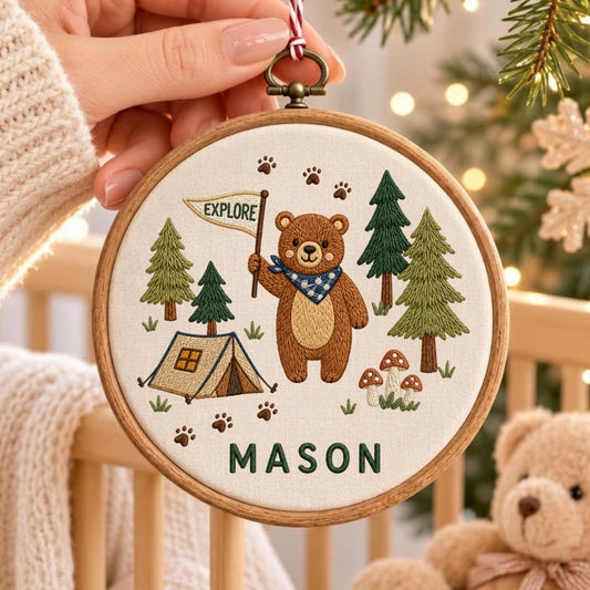 May include: A circular wooden embroidery hoop featuring a stitched design of a brown bear holding a flag that says EXPLORE. The scene includes green pine trees, a tan tent, mushrooms, and paw prints on cream fabric with the name MASON in green.