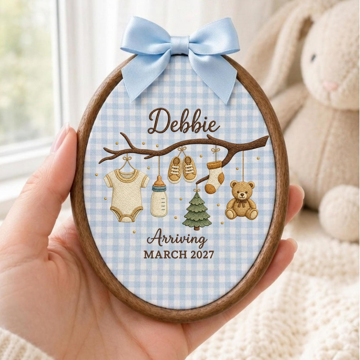 May include: An oval wooden embroidery hoop frame containing fabric with a light blue gingham pattern. It features a light blue satin bow at the top and an illustration of a branch with a onesie, baby bottle, shoes, sock, tree, and teddy bear. Text reads: Debbie Arriving MARCH 2027.