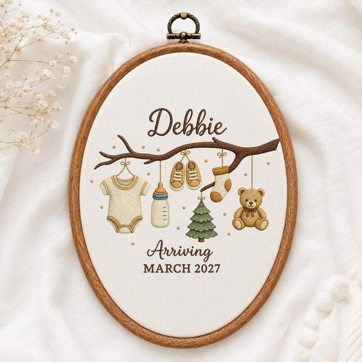May include: An oval wooden embroidery hoop containing white fabric with a brown embroidered branch. Hanging from the branch are a baby onesie, a bottle, two shoes, a sock, a green tree, and a teddy bear. Text reads: Debbie Arriving MARCH 2027.